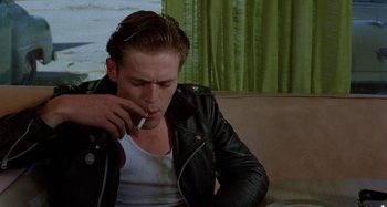 Movie still from “The Loveless” (1981), directed by Kathryn Bigelow – A man smoking a cigarette while sitting on a couch; Medium shot, Over the shoulder angle