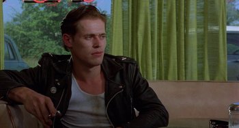 Movie still from “The Loveless” (1981), directed by Kathryn Bigelow – A man wearing a leather jacket sitting on a couch; Close Up shot, Over the shoulder angle