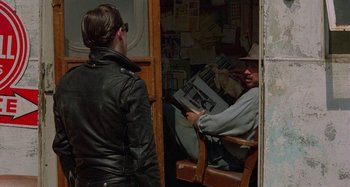 Movie still from “The Loveless” (1981), directed by Kathryn Bigelow – A man sitting in a chair reading a newspaper while a woman stands next to him; Medium shot, Over the shoulder angle