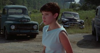 Movie still from “The Loveless” (1981), directed by Kathryn Bigelow – A young woman smoking a cigarette while standing next to old cars; Close Up shot, Over the shoulder angle