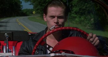 Movie still from “The Loveless” (1981), directed by Kathryn Bigelow – A man sitting in the driver's seat of a car smoking a cigarette; Close Up shot, Low angle
