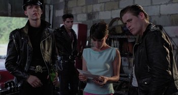 Movie still from “The Loveless” (1981), directed by Kathryn Bigelow – A woman standing next to a group of men; Medium shot, Over the shoulder angle