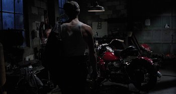 Movie still from “The Loveless” (1981), directed by Kathryn Bigelow – A man standing next to a red motorcycle in a garage; Wide shot, High angle