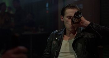 Movie still from “The Loveless” (1981), directed by Kathryn Bigelow – A man in a leather jacket drinking from a bottle; Medium shot, Over the shoulder angle