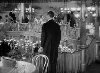 Movie still from “The Merry Widow” (1934), directed by Ernst Lubitsch – A black and white photo of a man standing in a restaurant; Wide shot, High angle