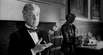 Movie still from “The Mirror Crack'd” (1980), directed by Guy Hamilton – A man in a suit and bow tie holding a cigarette; Medium shot, Over the shoulder angle