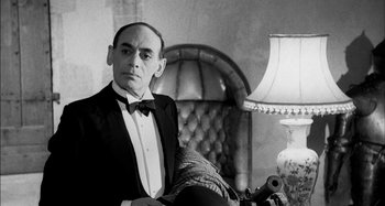 Movie still from “The Mirror Crack'd” (1980), directed by Guy Hamilton – A man in a tuxedo sitting in a chair in front of a lamp; Medium shot, Low angle