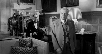 Movie still from “The Mirror Crack'd” (1980), directed by Guy Hamilton – A man in a suit standing next to another man in a suit; Medium shot, Low angle