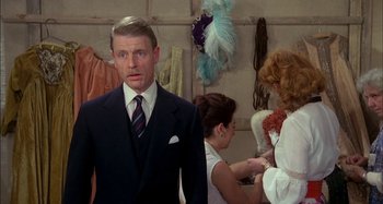 Movie still from “The Mirror Crack'd” (1980), directed by Guy Hamilton – A man wearing a suit and tie standing in front of a group of people; Medium shot, Over the shoulder angle