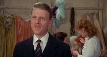 Movie still from “The Mirror Crack'd” (1980), directed by Guy Hamilton – A man wearing a suit and tie standing in front of other people; Close Up shot, Over the shoulder angle