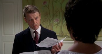 Movie still from “The Mirror Crack'd” (1980), directed by Guy Hamilton – A man in a suit and tie holding a piece of paper in front of a woman; Close Up shot, Over the shoulder angle