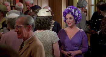 Movie still from “The Mirror Crack'd” (1980), directed by Guy Hamilton – A woman in a purple dress with flowers in her hair; Medium shot, Over the shoulder angle
