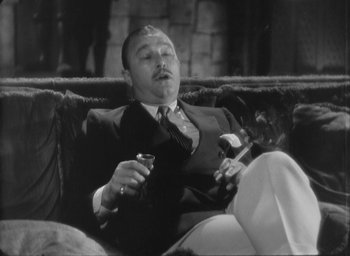 Movie still from “The Most Dangerous Game” (1932), directed by Irving Pichel – A black and white photo of a man smoking a cigarette and holding a glass of wine; Medium shot, High angle