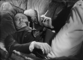 Movie still from “The Most Dangerous Game” (1932), directed by Irving Pichel – A black and white photo of a man smoking a cigarette and drinking a glass of wine; Medium shot, High angle