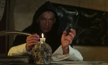 Movie still from “The Pit and the Pendulum” (1991), directed by Stuart Gordon – A man sitting at a table lighting a candle on top of a skull; Medium shot, High angle