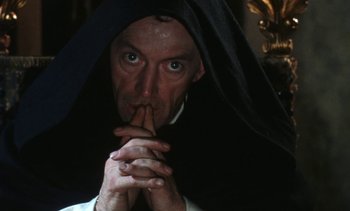Movie still from “The Pit and the Pendulum” (1991), directed by Stuart Gordon – A man in a priest's outfit with his finger to his mouth; Close Up shot, Low angle