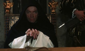 Movie still from “The Pit and the Pendulum” (1991), directed by Stuart Gordon – A man sitting at a table with his hands clasped together; Medium shot, High angle
