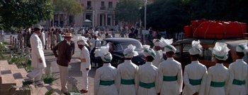 Movie still from “The Rains of Ranchipur” (1955), directed by Jean Negulesco – A group of men in white coats and hats standing in front of a car; Wide shot, High angle