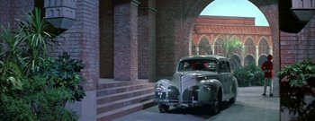 Movie still from “The Rains of Ranchipur” (1955), directed by Jean Negulesco – An old car parked in front of a brick building; Wide shot, High angle