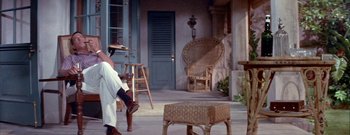 Movie still from “The Rains of Ranchipur” (1955), directed by Jean Negulesco – A person walking on a porch near a chair and a stool; Wide shot, High angle
