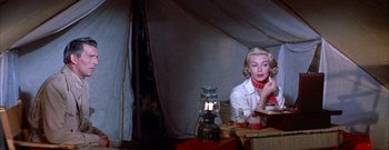 Movie still from “The Rains of Ranchipur” (1955), directed by Jean Negulesco – A woman sitting at a table in front of a lamp; Medium shot, Low angle