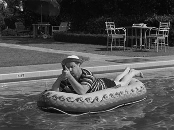 Movie still from “The Reluctant Dragon” (1941), directed by Jack Cutting – A man in striped shirt and hat floating on an inner tube in a swimming pool; Wide shot, High angle