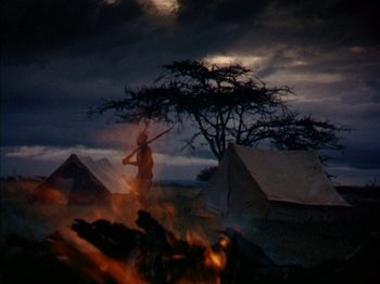 Movie still from “The Snows of Kilimanjaro” (1952), directed by Henry King – A person holding a stick in front of a fire; Wide shot, High angle