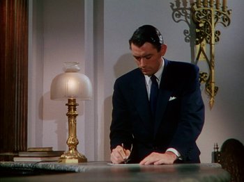 Movie still from “The Snows of Kilimanjaro” (1952), directed by Henry King – A man in a suit and tie writing on a piece of paper next to a lamp; Medium shot, High angle