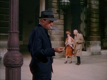 Movie still from “The Snows of Kilimanjaro” (1952), directed by Henry King – A man in a hat smoking a cigarette while standing next to a building; Wide shot, Over the shoulder angle