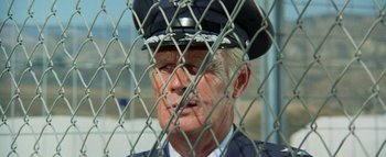Movie still from “The Swarm” (1978), directed by Irwin Allen – An older man in a police officer's uniform looks through a chain link fence; Close Up shot, Low angle