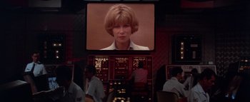 Movie still from “The Swarm” (1978), directed by Irwin Allen – A television screen showing a woman's face in front of people in a room; Medium shot, Low angle