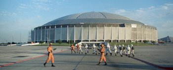 Movie still from “The Swarm” (1978), directed by Irwin Allen – A group of people in orange suits are walking in front of a building; Extreme Wide shot, Low angle