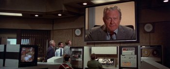 Movie still from “The Swarm” (1978), directed by Irwin Allen – A man in a suit and tie in front of a television; Medium shot, Over the shoulder angle