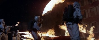 Movie still from “The Swarm” (1978), directed by Irwin Allen – A person in a spacesuit and a helmet standing in front of a fire; Wide shot, Low angle