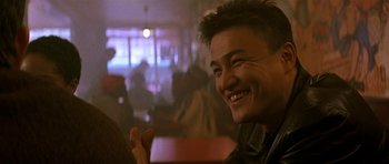 Movie still from “The Truth About Charlie” (2002), directed by Jonathan Demme – A man smiles while sitting at a table in a restaurant; Close Up shot, Over the shoulder angle