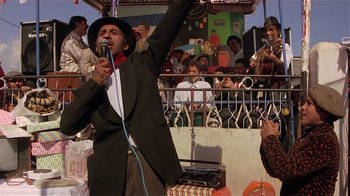 Movie still from “Time of the Gypsies” (1988), directed by Emir Kusturica – A man in a black jacket and a hat is holding a microphone; Medium shot, Low angle