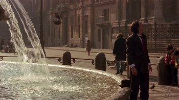 Movie still from “Time of the Gypsies” (1988), directed by Emir Kusturica – A man standing in front of a fountain on a city street; Wide shot, Low angle