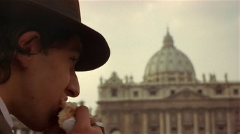 Movie still from “Time of the Gypsies” (1988), directed by Emir Kusturica – A man in a hat eating a sandwich in front of a building; Close Up shot, Over the shoulder angle