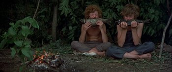 Movie still from “Tom Sawyer” (1973), directed by Don Taylor – A man sitting on the ground holding a fish in front of his face; Medium shot, Low angle
