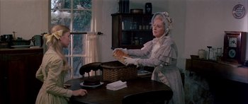 Movie still from “Tom Sawyer” (1973), directed by Don Taylor – An older woman in a white apron and a hat is putting a loaf of bread into a basket; Medium shot, Over the shoulder angle