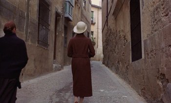 Movie still from “Tristana” (1970), directed by Luis Buñuel – A woman in a long coat walking down a street; Wide shot, High angle