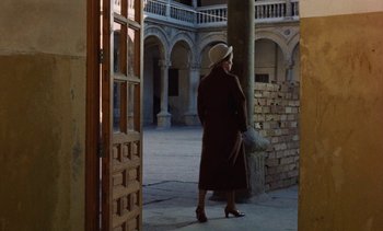 Movie still from “Tristana” (1970), directed by Luis Buñuel – An older woman in a hat and coat walking through a courtyard; Wide shot, Low angle
