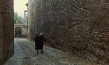 Movie still from “Tristana” (1970), directed by Luis Buñuel – Two people walking down a street next to a stone wall; Extreme Wide shot, High angle