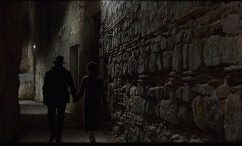 Movie still from “Tristana” (1970), directed by Luis Buñuel – A man and a woman holding hands in front of a stone wall; Wide shot, High angle