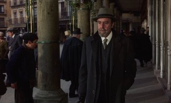 Movie still from “Tristana” (1970), directed by Luis Buñuel – A man in a hat and a coat standing next to a pillar; Medium shot, Over the shoulder angle