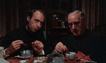 Movie still from “Tristana” (1970), directed by Luis Buñuel – Two men sitting at a dinner table with a plate of food in front of them; Close Up shot, High angle