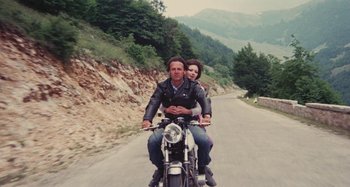 Movie still from “Your Vice Is a Locked Room and Only I Have the Key” (1972), directed by Sergio Martino – A man and a woman riding a motorcycle down a road; Wide shot, High angle