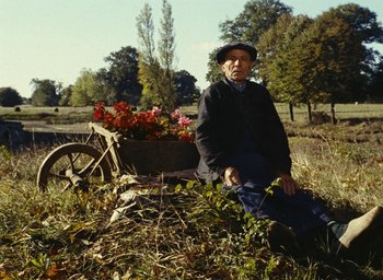 Movie still from “Ô saisons ô chateaux” (1958), directed by Agnès Varda – An old man sitting in the grass next to a cart of flowers; Wide shot, High angle