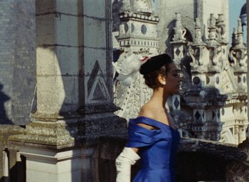 Movie still from “Ô saisons ô chateaux” (1958), directed by Agnès Varda – A woman in a blue dress walking in front of a building; Medium shot, Low angle
