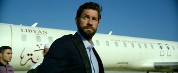 Movie still from “13 Hours” (2016), directed by Michael Bay – A man in a suit and tie standing in front of an airplane; Medium shot, Low angle